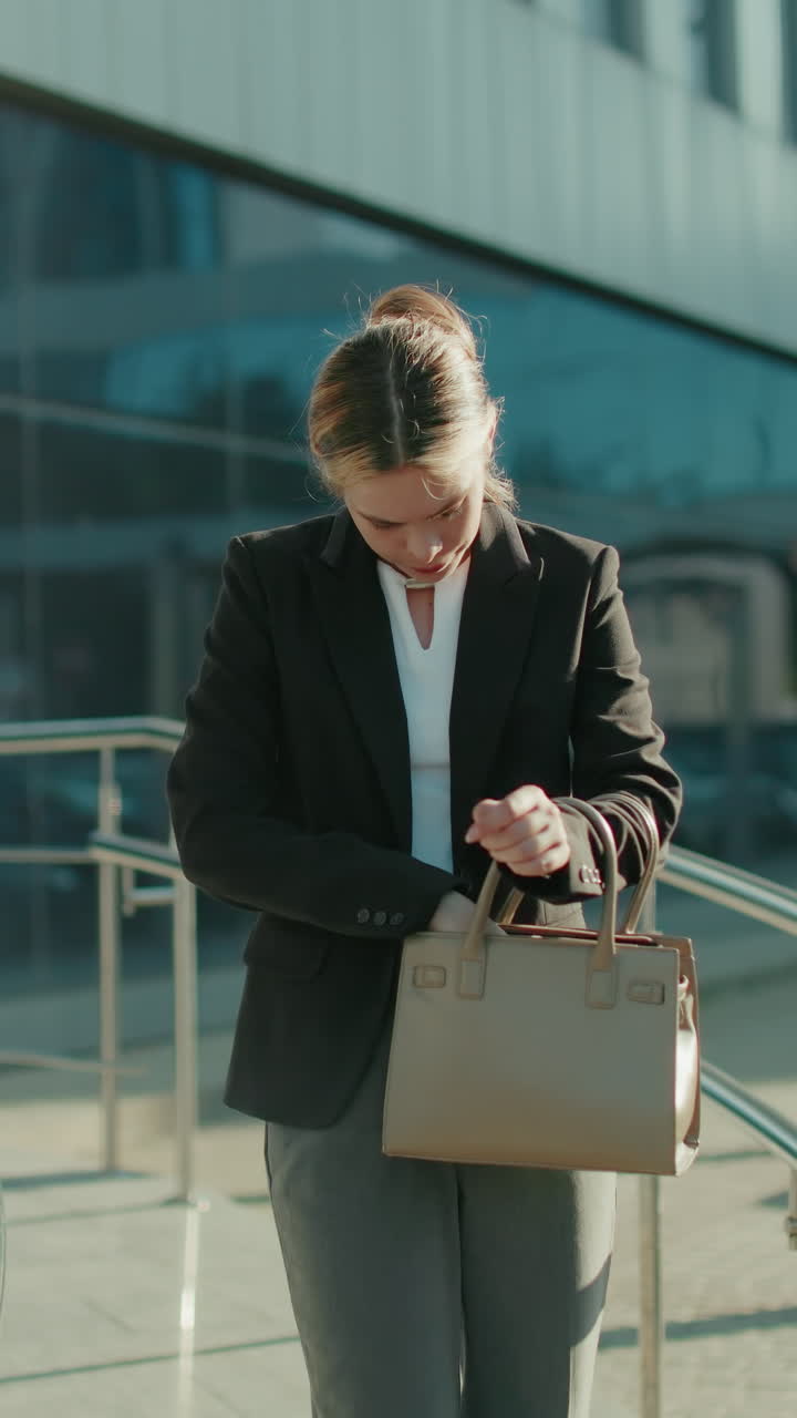 Professional woman walking down office ramp, contemplatively checking handbag to retrieve car key, surrounded by sleek metal railings, glass building reflecting urban setting