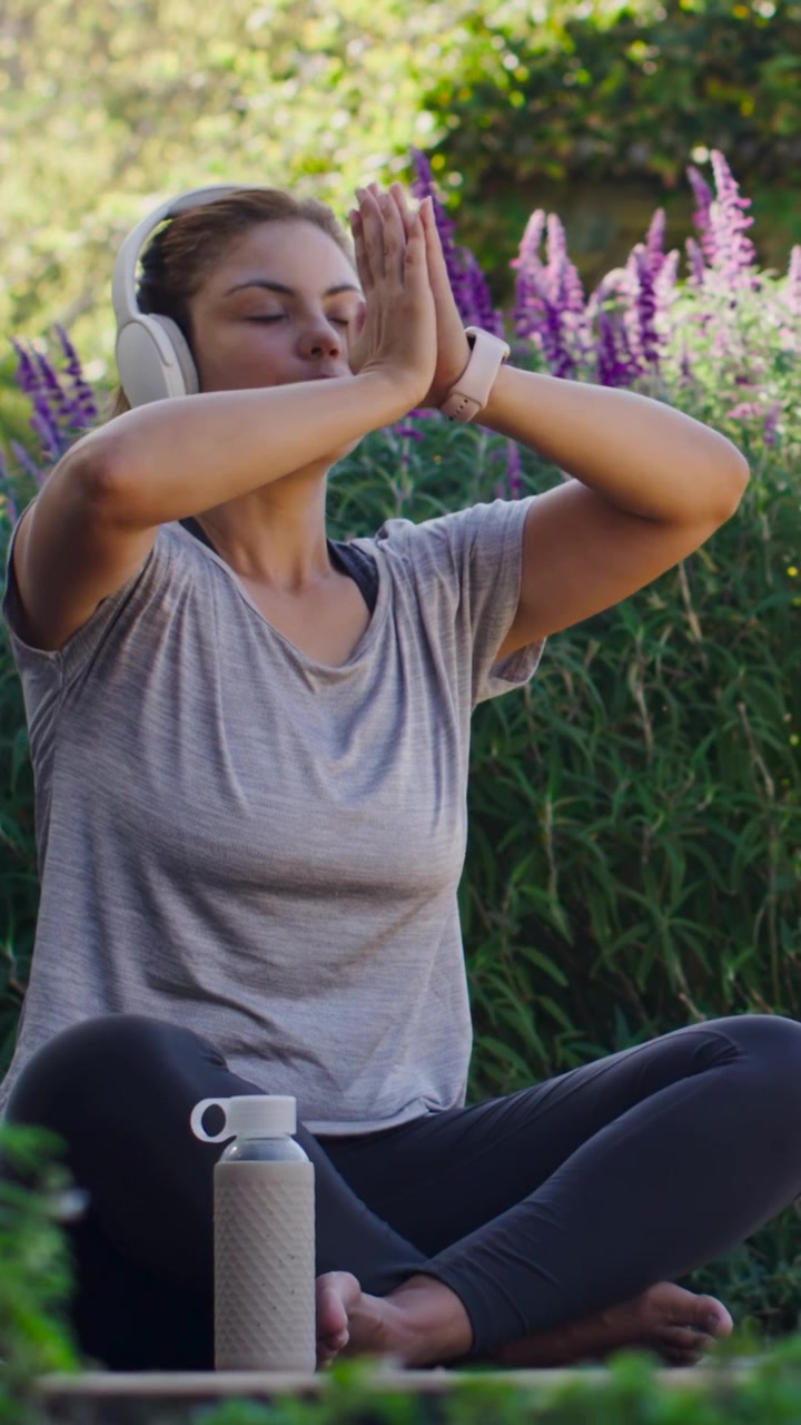 Video vertical: Después de inhalar, mujer con camiseta gris bajando las palmas para calmarse en el jardín con auriculares