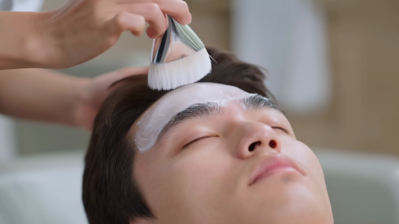 Asian Man Receiving Facial Brush Treatment In Serene Spa, Professional Esthetician Applying Creamy Mask