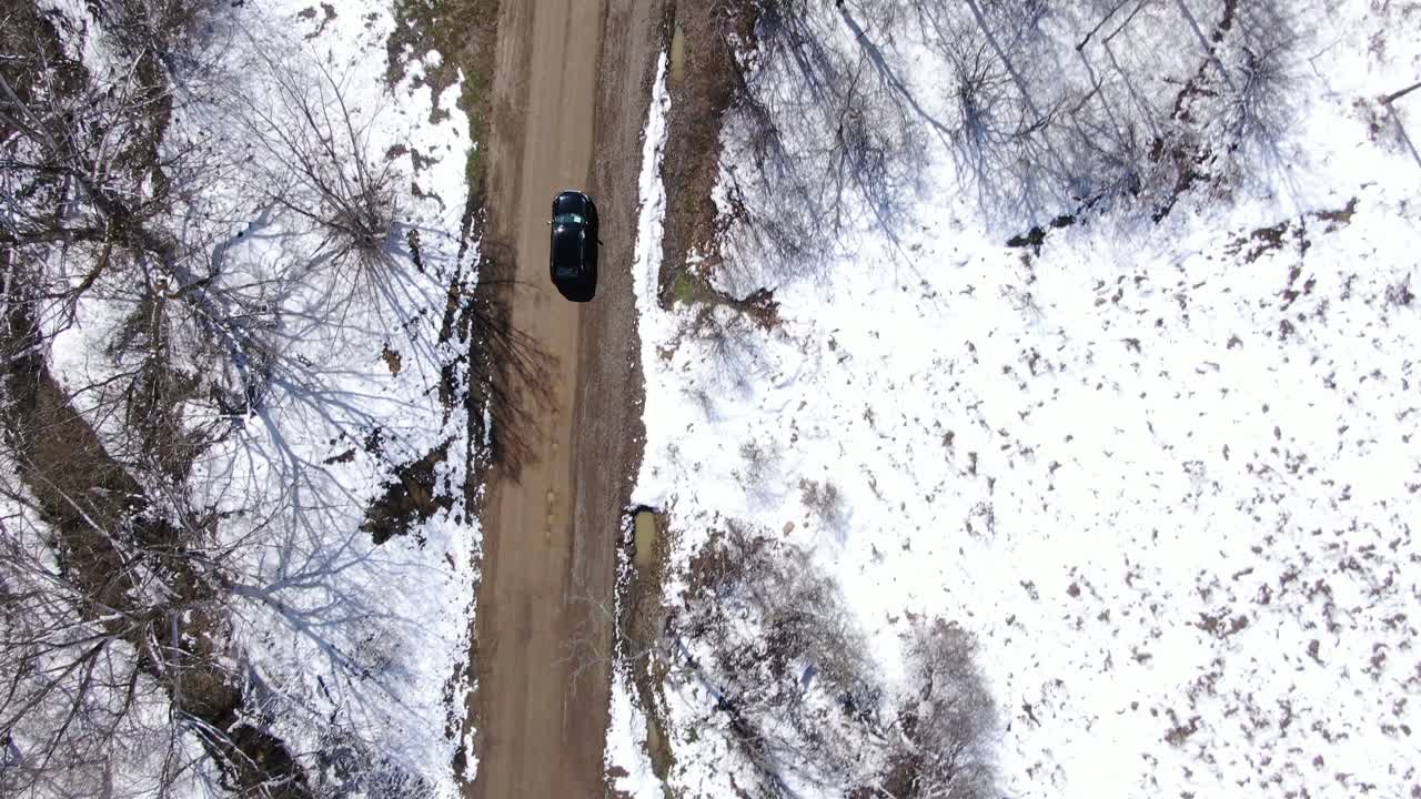 imágenes aéreas de drones de arriba hacia abajo de un sedán negro conduciendo por un camino de tierra en un bosque cubierto de nieve