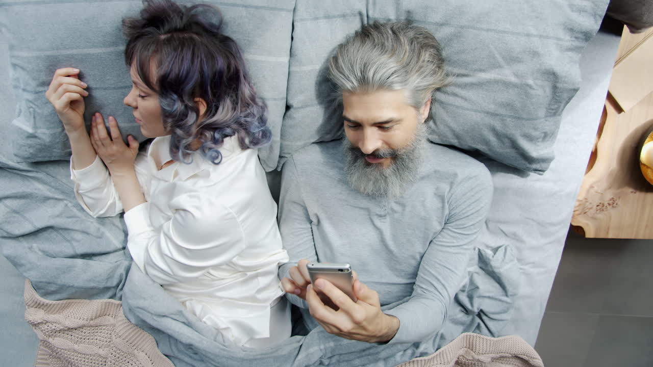 Couple in Bed, One Using Smartphone