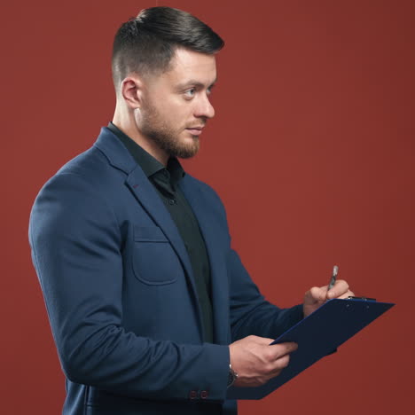 half-length video of handsome young smiling businessman wearing black shirt and dark blue suit holding the folder taking notes. dark red background.