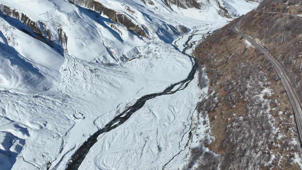 A winding river flows through a snow-covered valley, flanked by rugged slopes. The tranquil scene captures the essence of winter's calm and untouched beauty