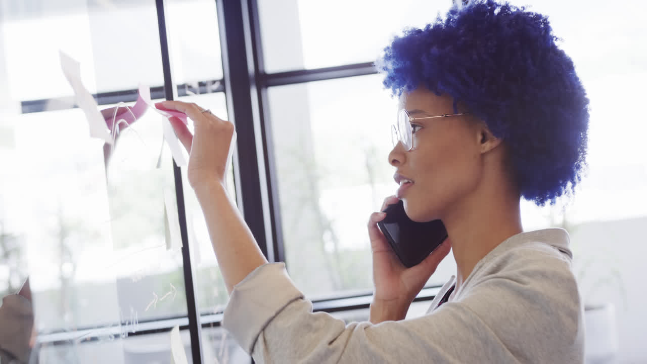 Biracial casual businesswoman making notes on glass wall and talking on smartphone, slow motion