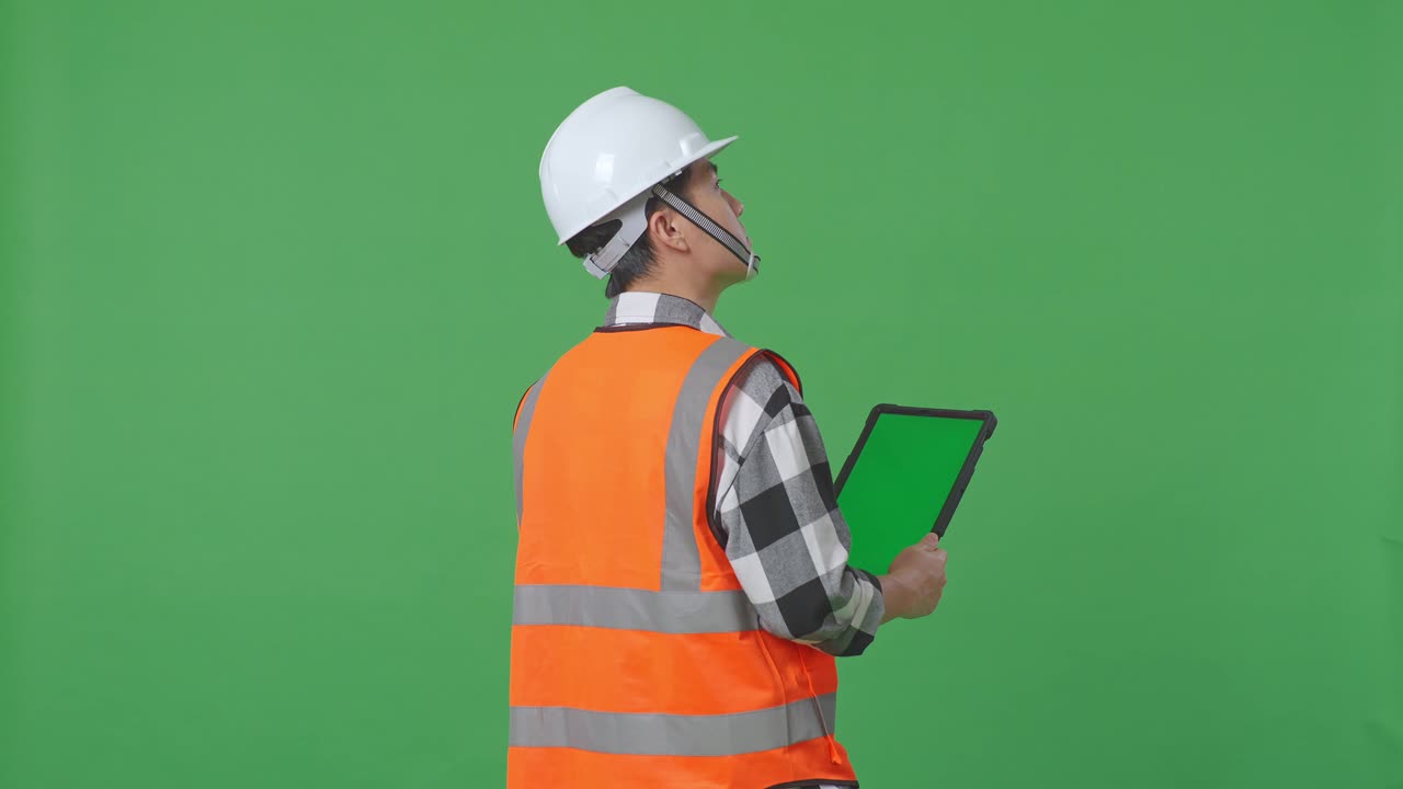 Back View Of Asian Male Engineer With Safety Helmet Working On A Green Screen Tablet And Looking Around While Standing In The Green Screen Background Studio