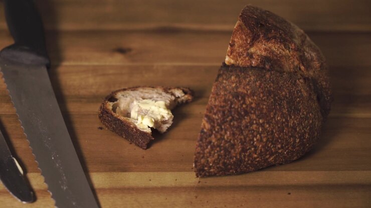 Static close up shot of a delicious and tasty slice of sourdough bread just after being cut, buttered then taken a bite out of and placed back on the board. Changing focus from the loaf to the slice.