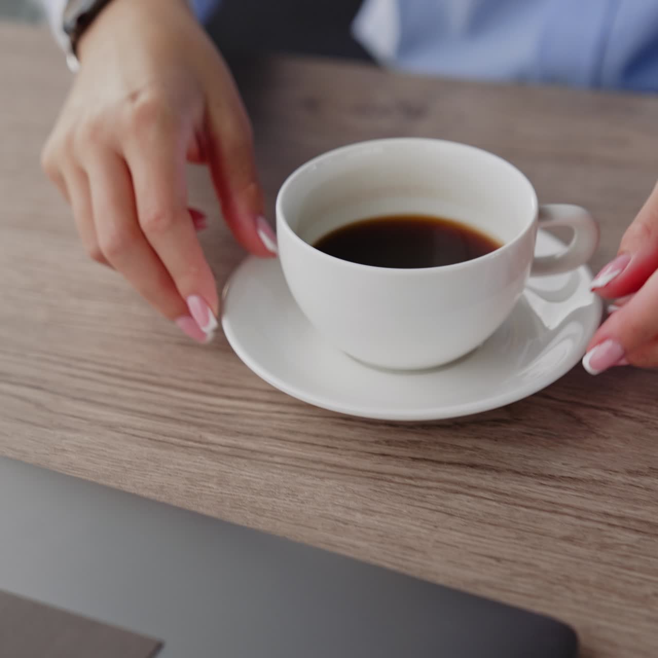 White cup of black coffee on the wooden table. Female hands pull the cup closer. Laptop and phone are on the desk beside. Close up