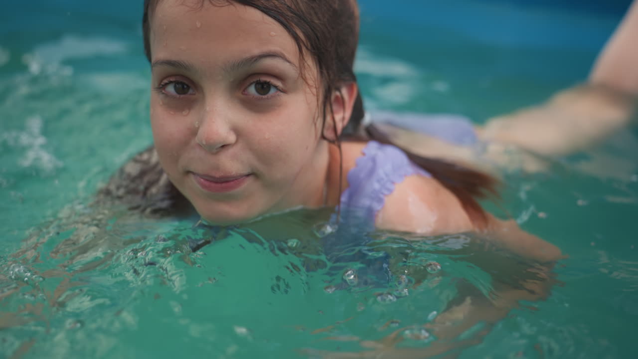Brunette Caucasian Girl In Backyard Pool Glides Near Yellow Inflatable Ball With Calm Smile, Steady Gaze And Wet Hair, Sunlight Reflections On Water, CloseUp Portrait Capturing Summer Leisure