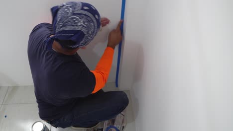 a man is employing blue masking tape to ensure precise and crisp lines while painting the wall - close up