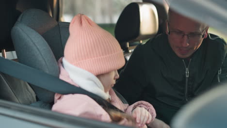 a man in a green jacket is carefully unbuckling the seat belt of his young daughter, who is dressed in a pink jacket and a matching pink beanie, inside a car