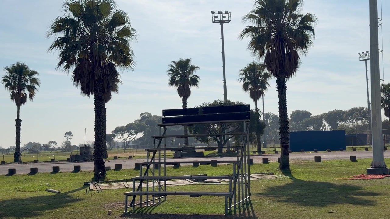 Static view of empty metal spectator bleachers at an outdoor sports field on a bright sunny day. Tall palm trees surround the ground