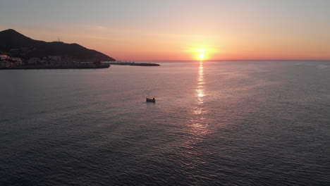 Sunrise-drone-shot-encircling-single-fishing-boat-surrounded-by-calm-sea-waves-near-coastline-town-with-mountain-range-backdrop