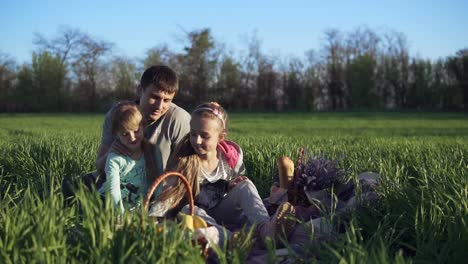 Glückliche-Familie,-Vater-Und-Töchter-Sitzen-Eng-Zusammen-Und-Genießen-Die-Zeit-Beim-Picknick-Auf-Der-Grünen-Schönen-Wiese-Auf