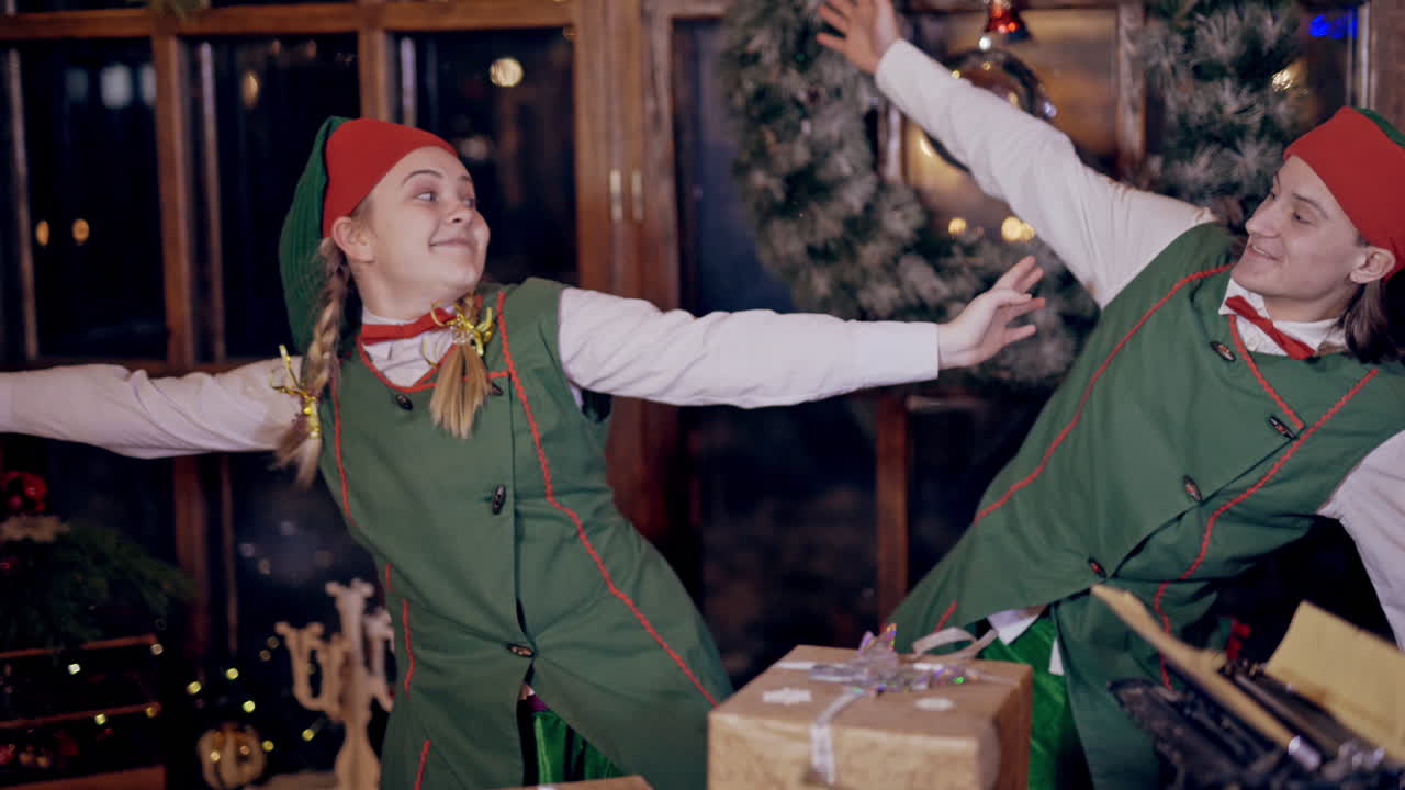 Cheerful elves in festive costumes imitate flying indoors. Happy man and woman in elf costumes are having a good time together in the room with decorations before Christmas.