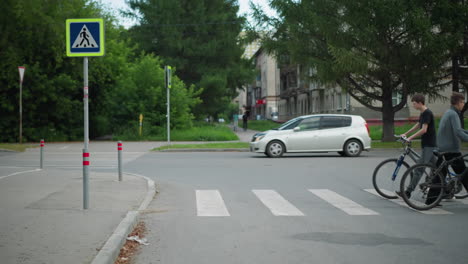 two people crossing a pedestrian crosswalk in opposite directions with a bicycle, a car is passing while a black car approaches, in the distance, with blur view of two people walking in the distance