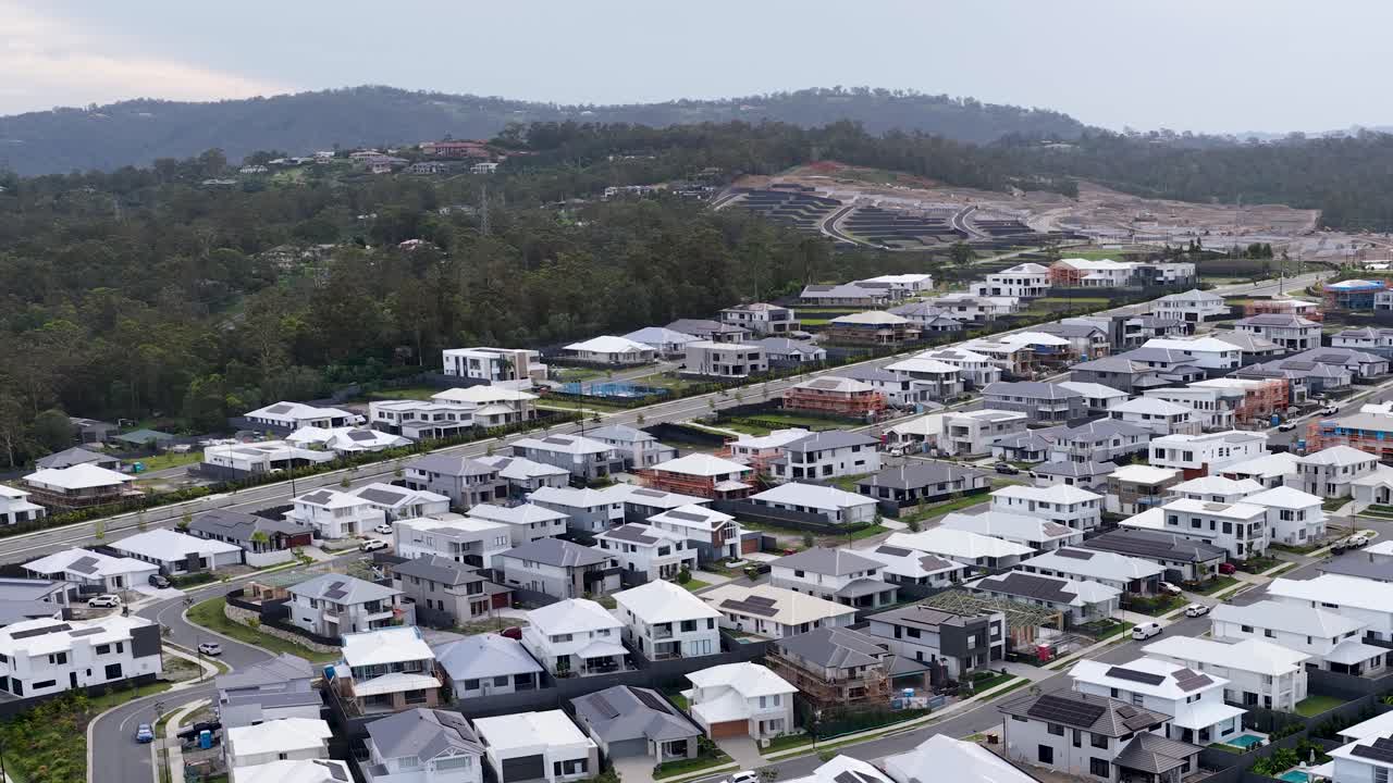 Drone slowly pans over contemporary suburban homes, lush forest, and distant mountains under soft daylight