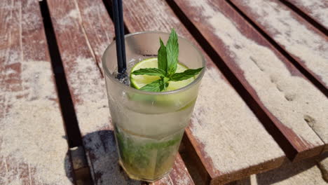 Ein-Glas-Mojito-Cocktail-Auf-Einem-Sandigen-Holzdeck-Am-Strand,-Nahaufnahme,-Drehen,-Drehen