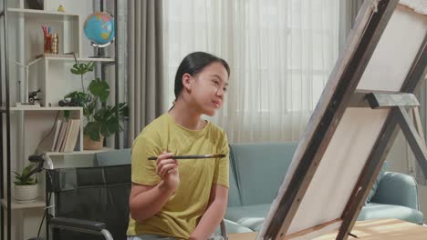 asian artist girl in wheelchair holding paintbrush thinking and painting on the canvas
