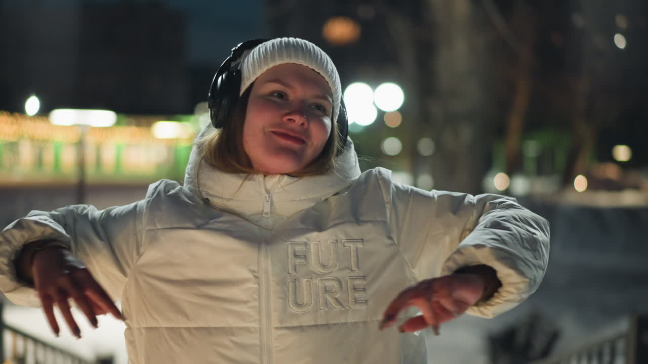 Close up front view of student in white winter coat and headphones smiling cheerfully as she sways hands and body to music in snowy park at night with ambient lights and faint people passing by