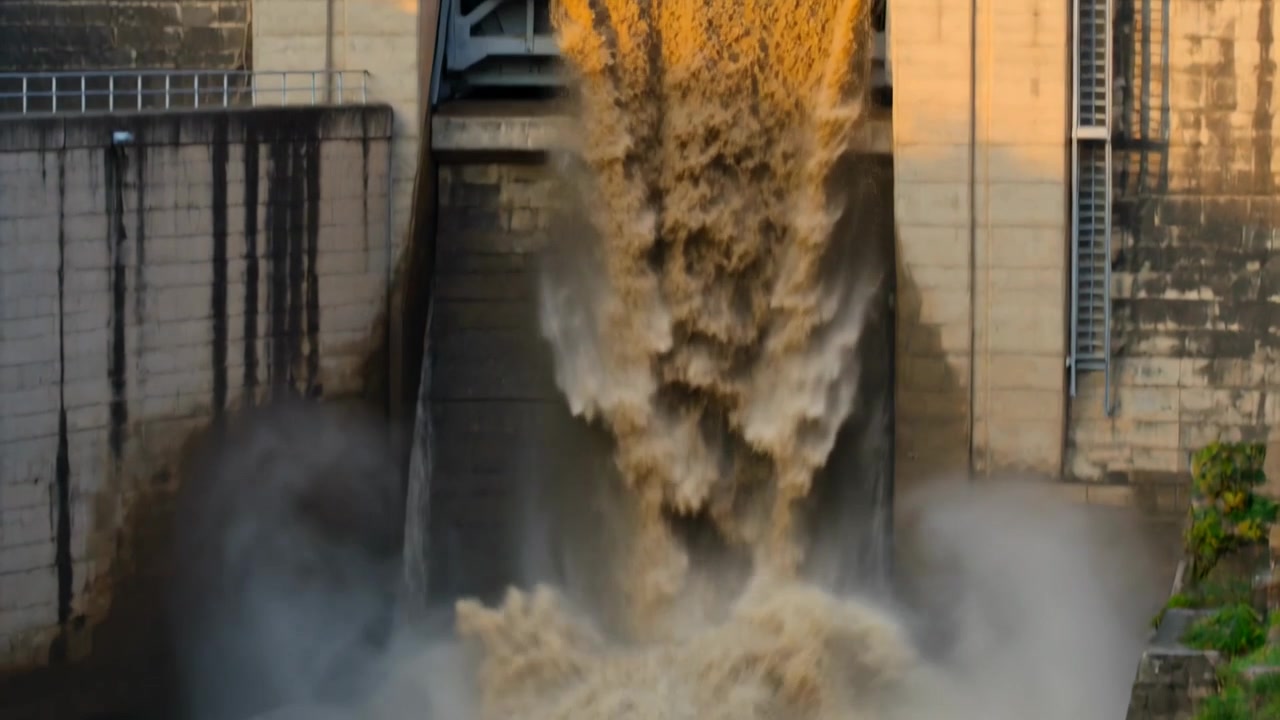Apertura di un cancello metallico, sfioratore che rilascia acqua fangosa lungo lo scivolo della diga, riempiendo il canale a valle.