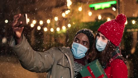 Nahaufnahme-Eines-Fröhlichen-Multiethnischen-Paares,-Das-Ein-Selfie-Mit-Dem-Smartphone-Macht,-Während-Es-Zu-Weihnachten-Auf-Der-Straße-Schneit