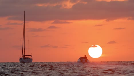 person sailing in motor boat toward sailboat and big sun on sunset above horizon at tropical destination, full frame