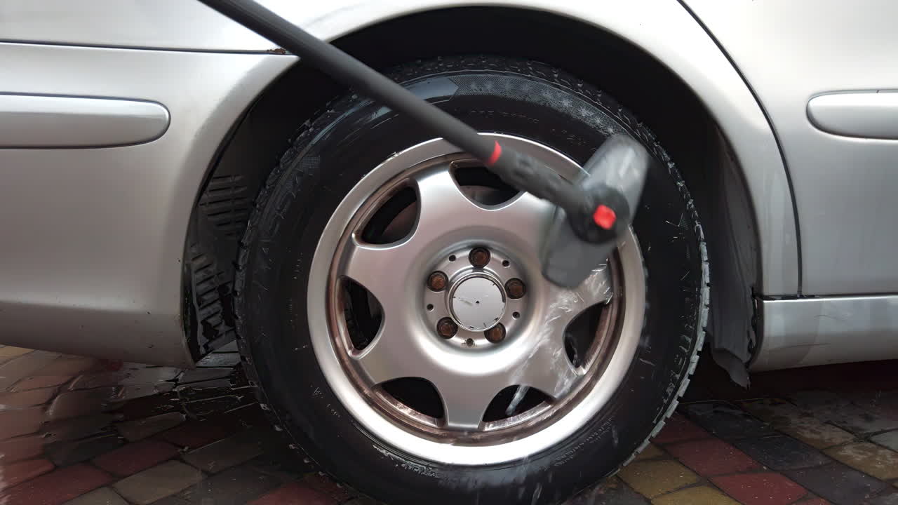 Bright afternoon in a driveway where an individual uses a pressure washer to clean the dirty tire of a silver car. Water and soap foam spray out as dirt washes away