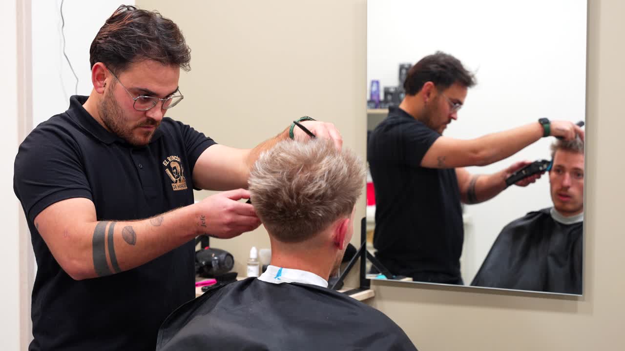 Fokuseret barber smiler til kunde, mens han klipper håret, set gennem spejlet