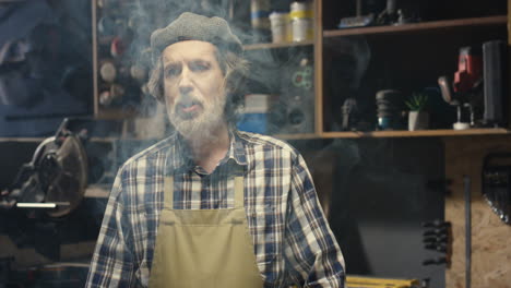 portrait of old caucasian handicraftsman in hat and apron smoking tobacco pipe while standing in carpentry workshop