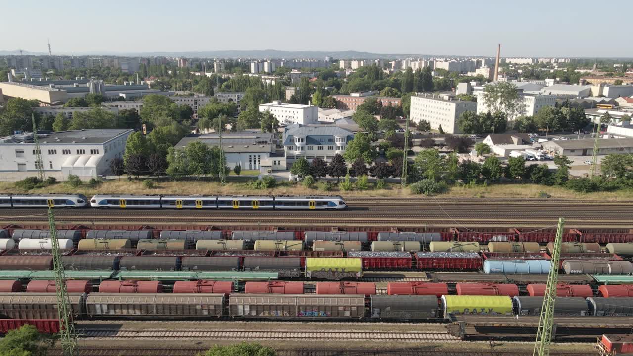 An aerial view of a busy railway station with multiple freight and passenger trains on parallel tracks. The background features an urban landscape with residential and industrial buildings.
