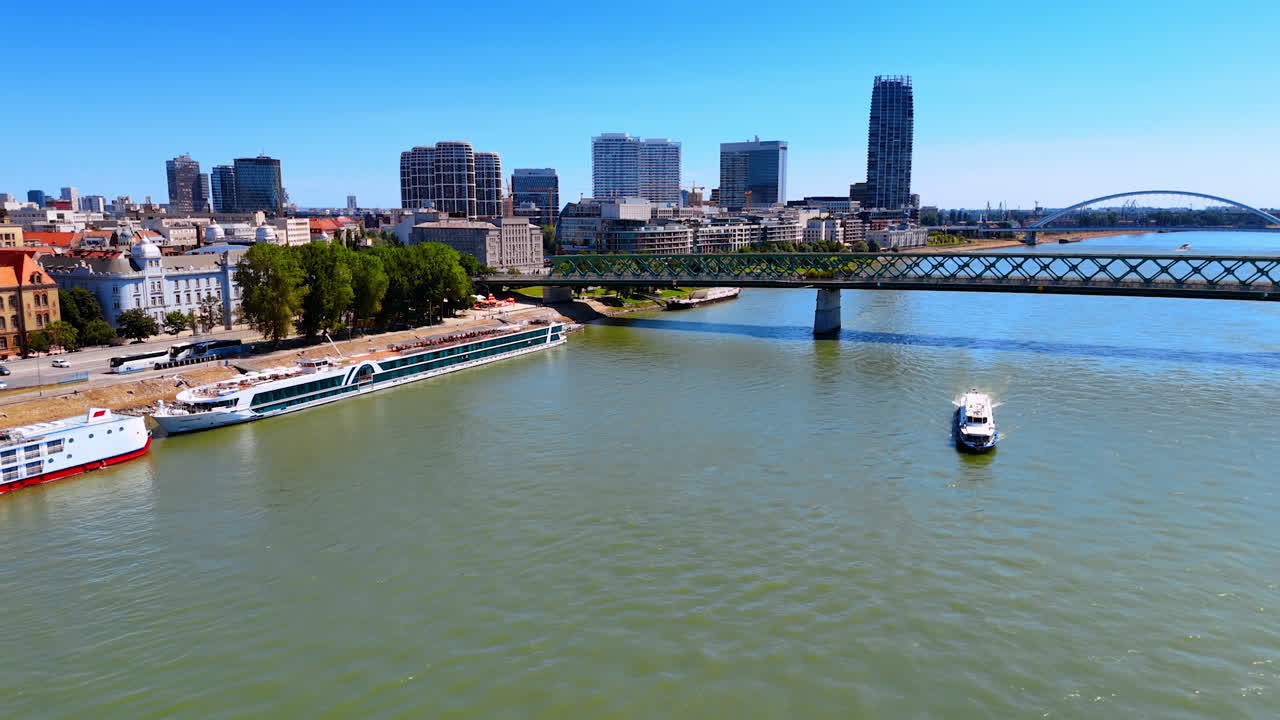 Long riverboat moves by the Danube River distancing from the Old Bridge. High-rises of Bratislava on the waterfront. Aerial view