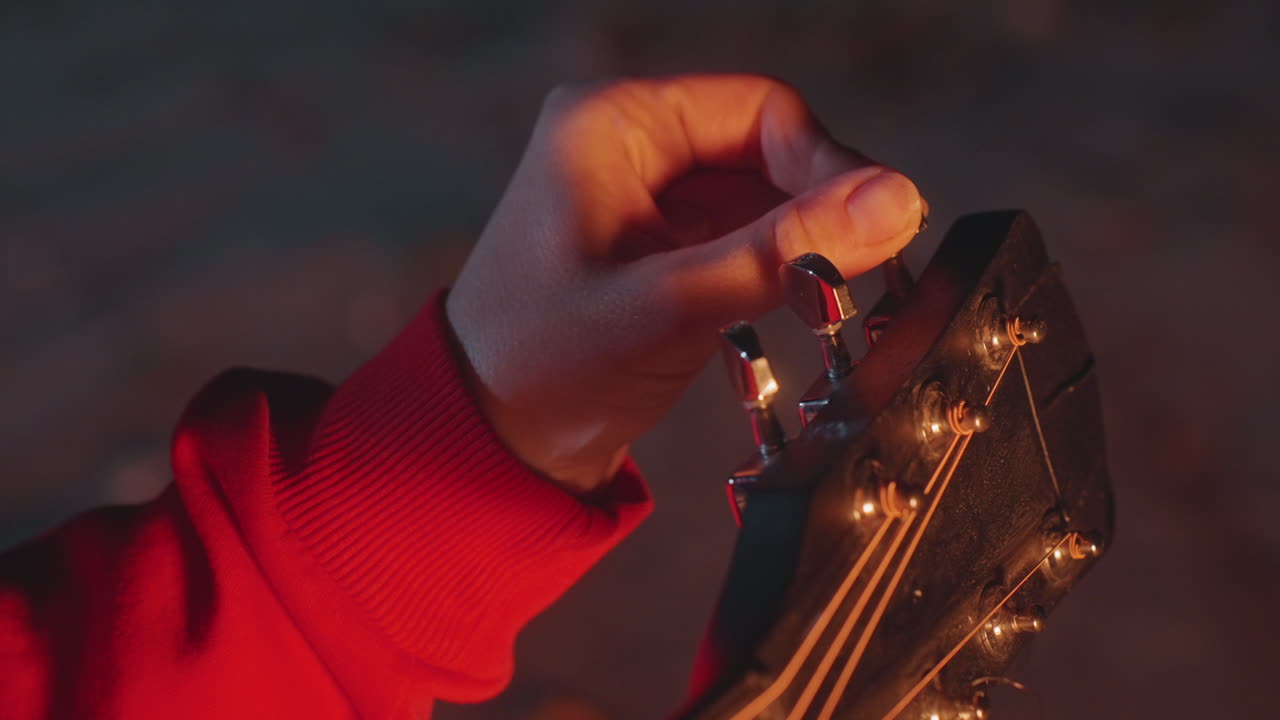 Campfire musician delicately tunes guitar strings. Farmer tunes guitar outside by warm glowing firelight. Musician finetunes their guitar outside under starlit sky near flickering campfire flame