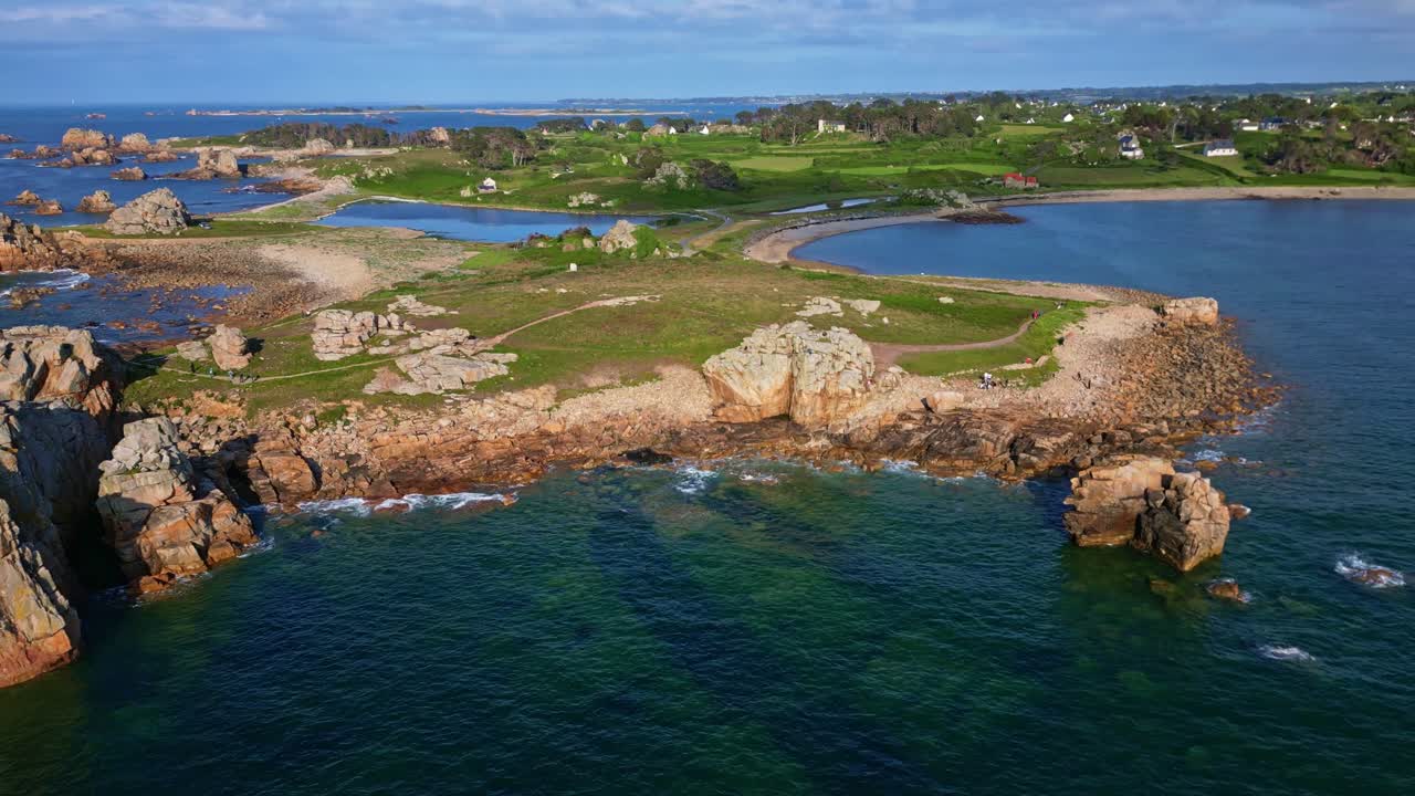Aerial drone pullback from Gouffre de Plougrescant showing rocky coast, hiking paths, vegetation, crashing waves, beach, and houses - Brittany France