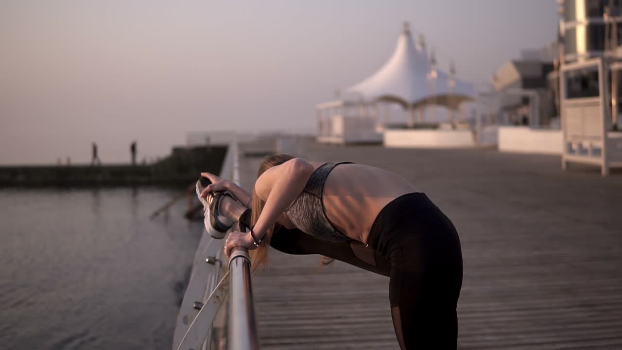 스포츠 의류 를 입은  ⁇ 고 적당 한 젊은 여성 이 바다 지역 에서 다리 근육 을  ⁇ 고 있다. 검은색 레 ⁇ 과 회색 브래지어를 입고 있다. 크로스바 에 의지 하고 있다. 측면 보기