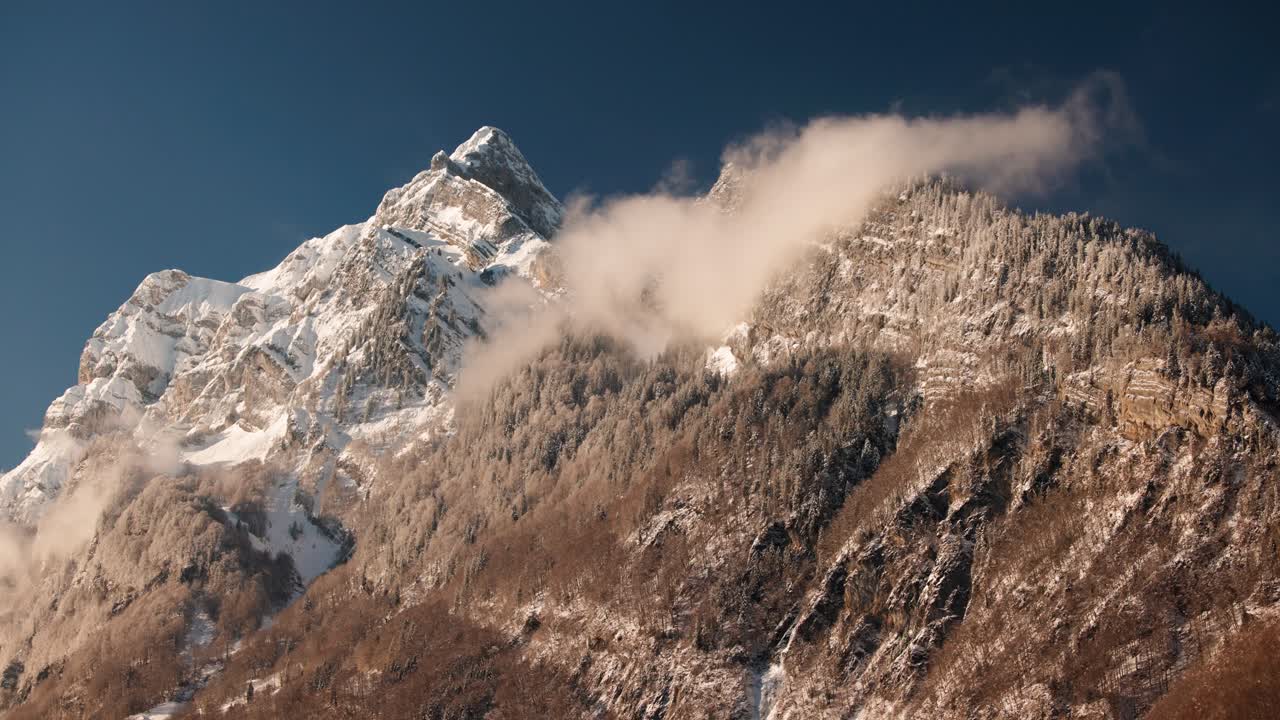 A telephoto timelapse of clouds moving quickly around a snow-covered alpine mountain. The static shot captures winter scenery, revealing shifting light and cloud motion under a clear blue sky