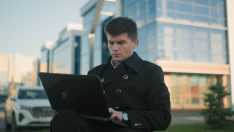 banquero sentado al aire libre operando una computadora portátil con las piernas cruzadas, expresión enfocada que muestra una intensa concentración, el fondo presenta edificios modernos de vidrio, coches estacionados y paisajes urbanos