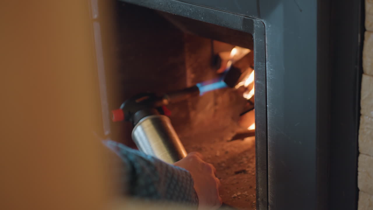 Close up of individual holding metal torch and directing blue flame into stacked firewood inside metal fireplace, fire begins to ignite slowly, creating warm glow
