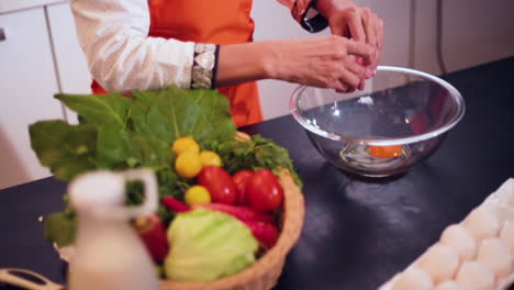 a slow mo close up view from top of a lady breaking an egg in the bowl in the kitchen, a basket of veggies with carrots, tomatoes and garlic on the table, the chef wore and orange colored apron