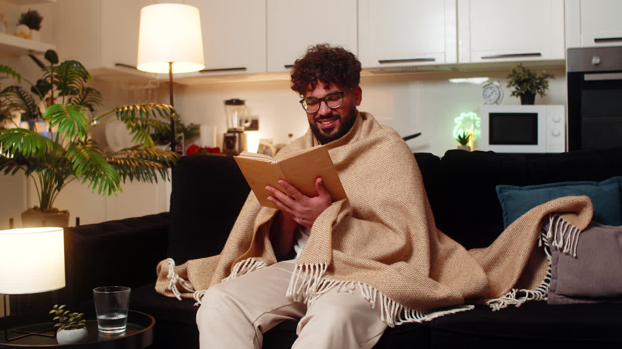 Arabian man in glasses wrapped in blanket reads novel on quiet evening relaxed mood focus calm peace