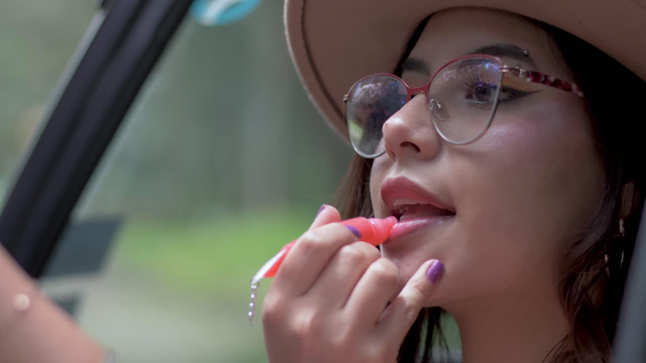 mujer elegante se aplica lápiz labial en los labios mirando el espejo del coche, adornada con sombrero y gafas, mostrando glamour y sofisticación en momentos cotidianos
