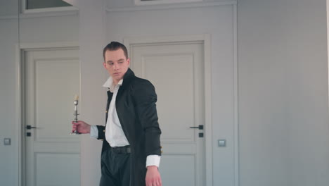 man holds candle stand while gazing at it stylishly in bright room with mirror reflecting apple on stool and lamp on stand showcasing confident pose and coat with subtle reflective white shirt detail