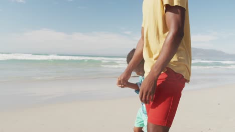 Afroamerikanischer-Sohn-Hält-Die-Hand-Seines-Vaters,-Während-Er-Am-Strand-Spaziert