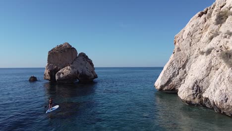 scenic view of a woman paddleboarding by large rocks in the mediterranean sea