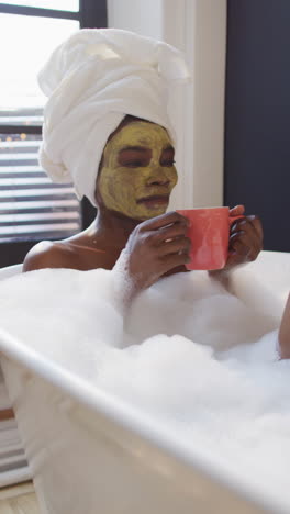 video vertical de una mujer afroamericana sonriente bebiendo café, mimándose en un baño de espuma