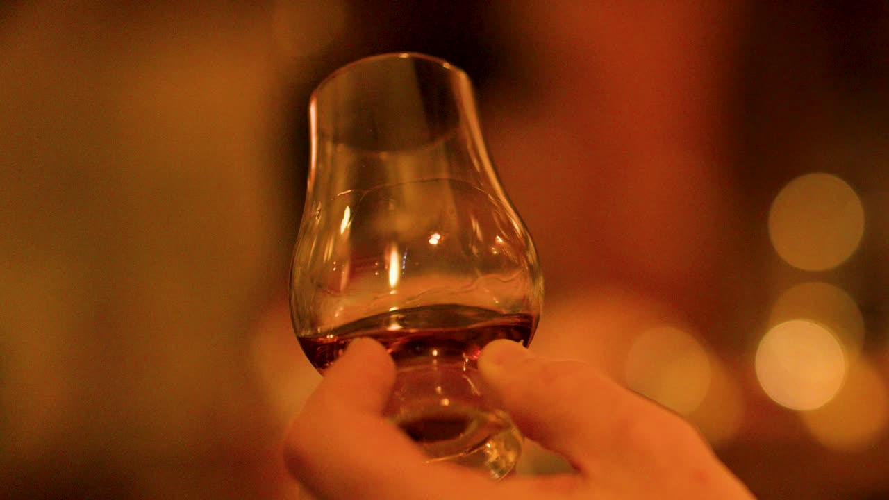 Hand swirls whisky glass in cozy bar, amber drink illuminated by soft, ambient evening lighting