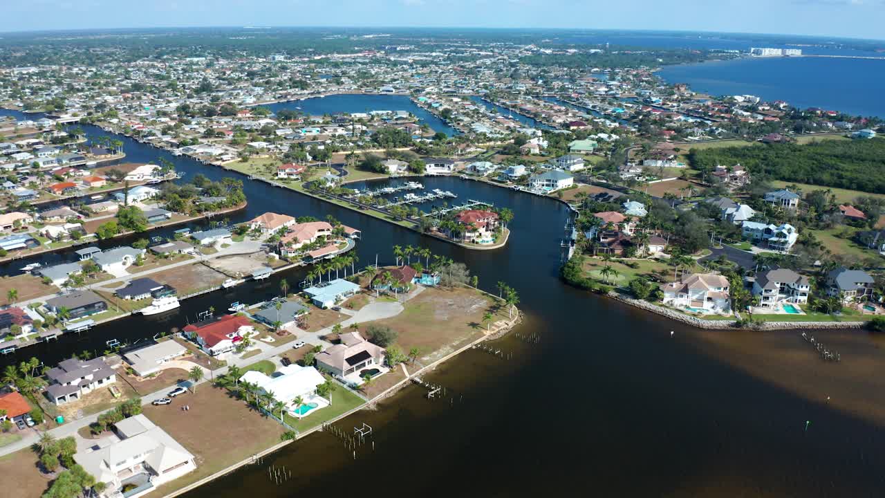 A coastal residential area in Florida featuring canals, boat docks, and numerous houses with landscaped yards adjacent to calm water and a distant shoreline under a clear blue sky