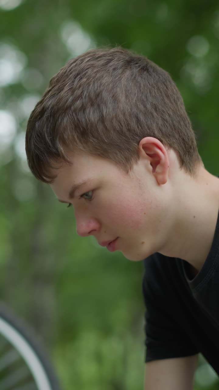 Young boy in black top sits thoughtfully on a park bench with his head down, next to his upside-down bicycle with slowly rotating tires, the serene background includes blurred trees
