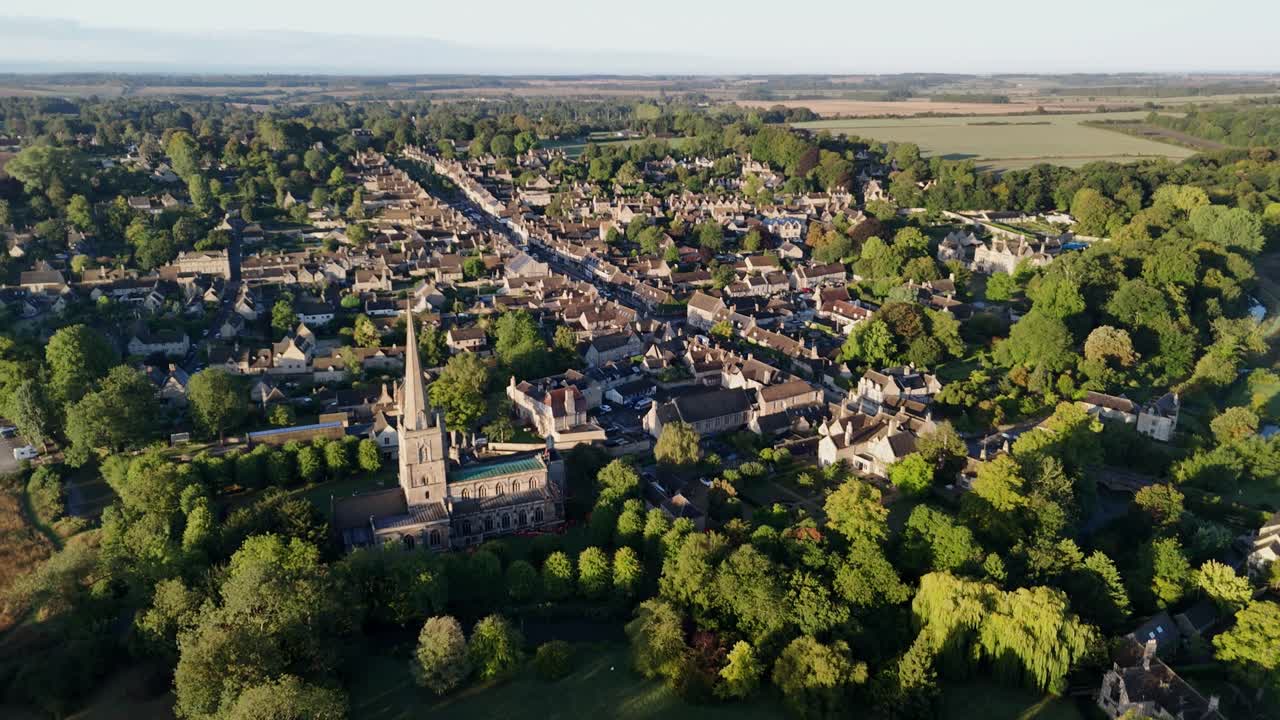 aerial drone flyover of the village of Burford in the Cotswolds, the historic High Street and golden stone buildings, bathed in the early morning sunlight