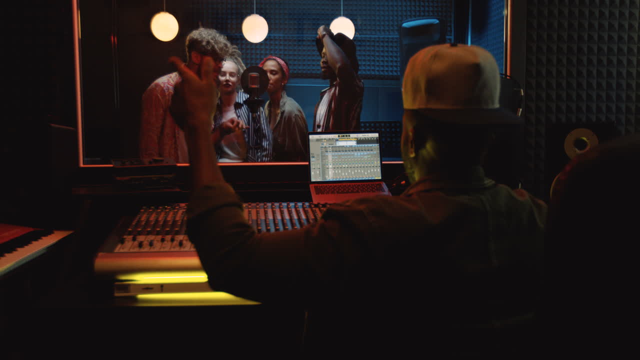 vista posterior del productor de sonido masculino sentado en el mezclador en el estudio oscuro y trabajando sobre la canción mientras la gente canta y graba en el micrófono