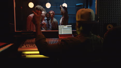 vista posterior del productor de sonido masculino sentado en el mezclador en el estudio oscuro y trabajando sobre la canción mientras la gente canta y graba en el micrófono